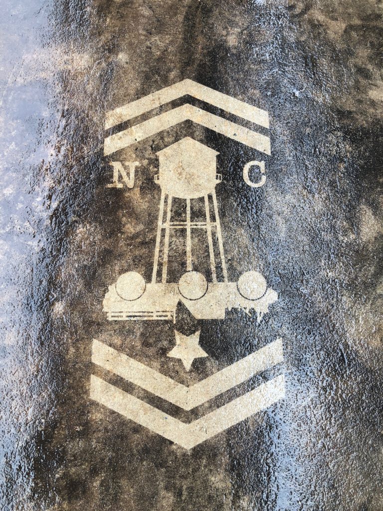 dirty sidewalk with pressure washed image of a water tower, camp north end logo and army arrows.