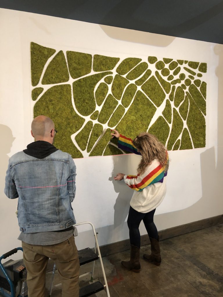 blonde woman, the founder of #thesavageway, installs the final moss piece of an installation.