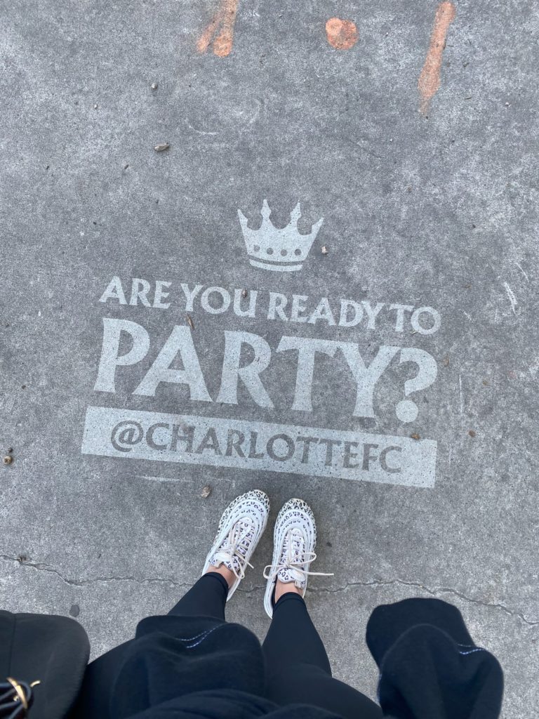 Photo of a feet standing near a Clean Graffiti message that reads "Are you ready to Party? @CharlotteFC" on a sidewalk in Charlotte, NC.